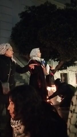 The video captures a nighttime scene outdoors, likely in Ñuñoa, Chile, given the visible text referencing "ESTADIO NACIONAL" and "AV. GRECIA". Several individuals are gathered, illuminated by artificial light.

In the foreground, a person wearing a black jacket and a light-colored, patterned head covering (possibly a keffiyeh) holds their right hand aloft with one finger extended. They have a dark bag slung over their shoulder. Next to them, a large white banner is being held by multiple people. Clearly visible text on the banner includes "AV. GRECIA" and "REPUBLICA DE ISRAEL," along with a downward-pointing arrow. Another banner, partially visible on the left, shows the words "ESTADIO NACIONAL."

Further back, other individuals are present, and a large tree with dark foliage stands on the left. The overall atmosphere suggests a gathering, possibly a protest or demonstration, indicated by the banners and the hand gesture. The lighting is dim, characteristic of an evening or night setting.