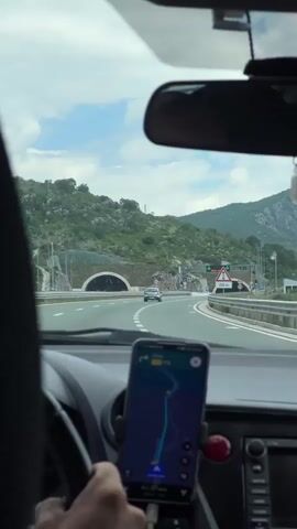 A first-person perspective shot from inside a car traveling on a highway. The driver's hands are visible on the steering wheel and a smartphone is mounted on the dashboard, displaying a navigation app. Ahead, the highway leads towards two tunnels nestled in a mountainous, green landscape. A few other cars are visible on the road. Signage near the tunnels indicates "TUNEL PLOCET," and another sign reads "200m." The sky is partly cloudy.