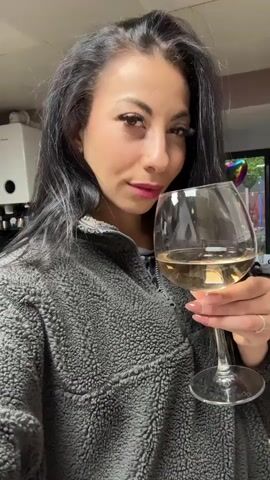 A woman with long dark hair, wearing a gray fleece jacket and pink lipstick, looks at the camera while holding a glass of white wine. She then raises the glass to her lips and takes a sip. The background includes a kitchen with white cabinets, a coffee machine, and a view of a backyard through a window. A heart-shaped balloon is visible in the yard.