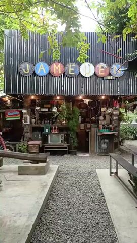 A vertical video shows the facade of a rustic shop with the word "JAMEUEN" displayed across the top in individual letter signs. The shop is adorned with various vintage items, including an old television, license plates, and potted plants. The area in front of the shop is covered in gravel, with concrete pathways and wooden benches, suggesting an outdoor seating area. Lush green foliage overhangs the scene from above, creating a natural and somewhat secluded atmosphere. The lighting is warm, with visible bulbs illuminating the shop's interior and exterior.