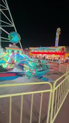 The video displays a nighttime scene at an amusement park. In the foreground, a yellow metal fence encloses a walkway. Beyond the fence, a teal and blue airplane-shaped ride is visible, rotating with its propeller in motion. Colorful lights illuminate the ride. In the midground, a game booth with a sign reading "MAGIC GAMES" is set up, with people browsing the games and staff behind the counter. Further in the background, a tall, illuminated tower ride with colorful lights stands against the dark night sky. Other amusement park rides and structures are also present, along with a Ferris wheel on the left side of the frame. Several people are walking around the park. The overall atmosphere suggests a lively carnival or fair.