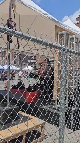 A concert is being set up under a white tent with a chain-link fence in the foreground. Two people are visible on a stage, one appears to be setting up equipment. There are red buckets and speakers on the stage. Other tents and buildings are visible in the background.