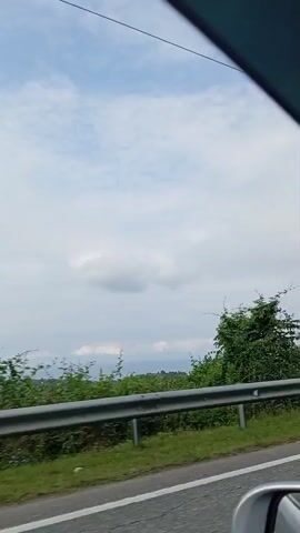 The video shows a view from a vehicle traveling along a road. The camera is looking out the side window, capturing the passing scenery. The foreground features a guardrail and green grass along the roadside. Beyond the guardrail are lush green trees and vegetation. The sky above is partly cloudy with a light blue hue. The overall impression is of a scenic drive through a natural landscape.