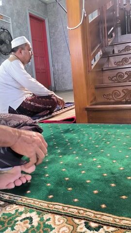 This video captures an indoor scene, likely within a mosque or prayer room, during daytime in Blangbitra, Indonesia. The main subject is a man performing Islamic prayer (Salah). He is wearing a white cap, a white long-sleeved shirt, and a brown patterned sarong, and is seen bowing in the *ruku'* position. Another individual, partially visible, is praying behind him.

In the mid-ground, a grand, multi-tiered wooden minbar or pulpit stands prominently, intricately carved with elaborate floral and swirling designs. The foreground is dominated by a vibrant green prayer rug adorned with small, evenly spaced gold diamond-shaped patterns. In the background, a bright red door is visible, along with a microphone on a stand, suggesting its use for announcements or sermons. The lighting is bright, illuminating the space. The audio features the Islamic declaration "Allahu Akbar" (God is the greatest), which is commonly recited during prayer. The scene conveys a sense of devotion and religious observance. No discernible text is visible within the frame.