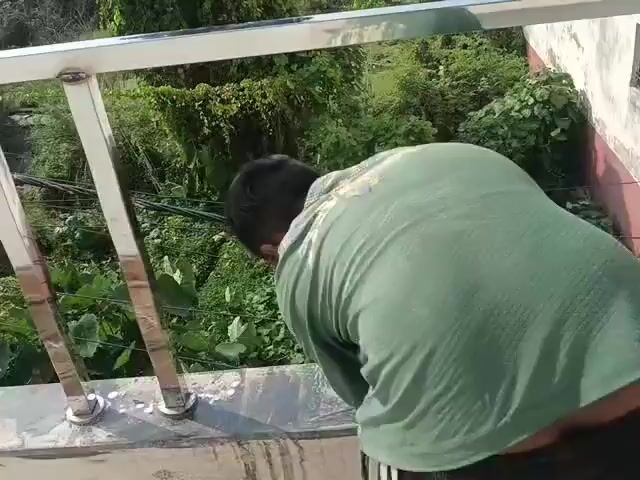 The video shows a person on a rooftop, working on a metal railing. The person is wearing a light-colored t-shirt and dark pants, and appears to be using a power tool to grind or polish the railing. The railing is made of stainless steel and has vertical supports. In the background, there are trees and vegetation, indicating an outdoor setting. The railing is attached to a concrete ledge or balcony. The person is positioned with their back to the camera and is focused on their work.