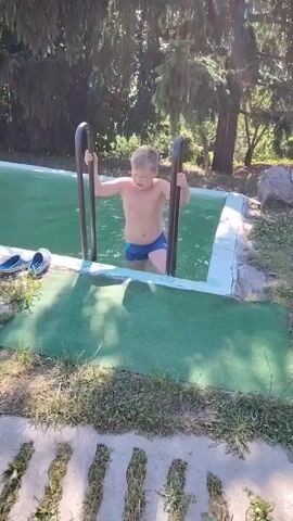 A young boy wearing blue swim trunks stands in a shallow swimming pool, holding onto the metal rails of a ladder. He is positioned at the edge of the pool, which has green carpeting around it. Blue and white shoes are visible on the pool deck to his left. The pool is surrounded by trees and foliage, suggesting an outdoor setting. The boy appears to be in the water up to his waist.