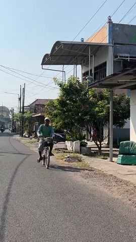 A man wearing a light blue shirt and white hat rides a bicycle down a paved road. The man is facing the camera and appears to be moving towards it. The bicycle has a basket on the front and a rack on the back. The man is looking straight ahead. The road is lined with buildings and trees. The sky is clear and blue. The sun is shining. There are power lines visible in the background.