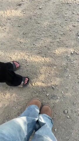 The video begins with a close-up shot of two pairs of feet standing on a dusty, gravelly path. The person on the left wears black pants and sandals, while the person on the right wears blue jeans and light brown boots. The camera then pans upwards and moves forward, revealing a winding dirt road through a hilly, wooded landscape. Telephone poles and power lines are visible along the side of the road. The sun appears to be setting, casting long shadows and illuminating the scene with a warm, golden light. The trees are lush and green, suggesting a temperate climate. The overall impression is one of a peaceful, outdoor setting, possibly during a hike or leisurely walk.