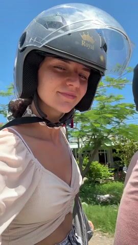 A woman with dark curly hair wears a gray helmet with a clear visor and a tan crop top. She is on the back of a motorcycle with two other people, one in front of her wearing a pink t-shirt and another person behind her wearing a black helmet. The motorcycle is moving along a paved road in a rural area under a clear blue sky. The woman turns her head to look at the camera and smiles slightly. Buildings and greenery are visible in the background.