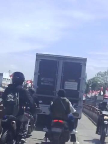 The video shows a view from behind motorcycles stopped on a bridge, waiting to move.  A truck blocks the view ahead. Indonesian flags line the bridge's railing. The bridge appears to be steel. The motorcycles and the truck are waiting in line. It is daytime.