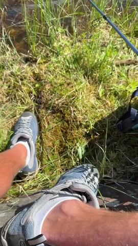 The video begins with a close-up of a person's legs and feet wearing athletic shoes and shorts, sitting by the edge of a body of water. A fishing rod is visible, extended over the water. The camera then pans upwards, revealing a wider view of the lake, surrounded by green grass and trees, with mountains in the distance. The water is rippling gently, and the sun is shining, casting light on the scene. The video captures a peaceful moment of someone fishing in a natural setting.