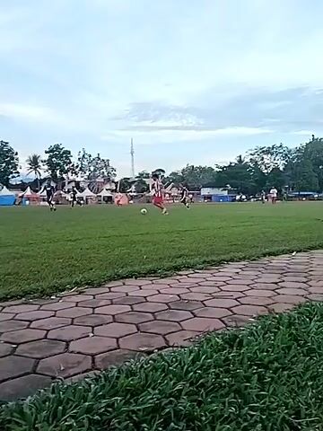 The video captures an outdoor soccer match or practice taking place on a green grass field. Several players, predominantly male, are actively engaged with the soccer ball. One player in a red and white striped jersey is prominently featured, dribbling the ball down the field.

In the background, a series of colorful tents (white, red, and blue) are set up, suggesting a local market, festival, or community event adjacent to the field. Beyond these tents, various trees, including palm trees, and a tall antenna or tower are visible. Distant mountains can be seen on the horizon under a partly cloudy sky.

The foreground of the shot features a paved pathway constructed from hexagonal blocks, bordering the playing field, with some low green foliage. The lighting suggests it is late afternoon or early evening, with a clear yet soft natural light. The overall scene depicts a lively community gathering centered around a sports activity, likely in Matesih, Indonesia. No visible text is discernible in the video.