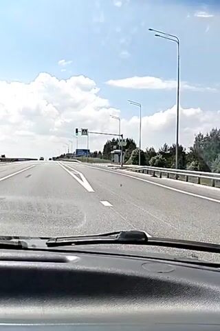 The video is filmed from the perspective of someone driving a car on a multi-lane highway. The road ahead is clear with white lines marking the lanes. There are streetlights and some trees on the right side of the road. A pedestrian crossing with yellow and white stripes is visible. Traffic lights are displayed, and one of them is green. There are signs on the right side of the road, including a bus stop shelter. The sky is blue with white clouds, indicating daytime. There are other cars visible in the distance. The dashboard and windshield wipers of the car are in the foreground.