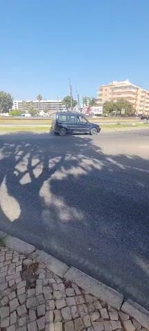 A blue van drives from left to right across the screen. A black car drives from right to left across the screen. A white van drives from left to right across the screen. The video is filmed on a sunny day with a clear blue sky. There are trees casting shadows on the road. In the background, there are buildings and a fountain.