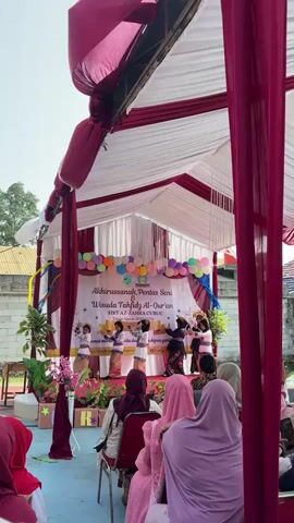 A group of children, dressed in what appear to be traditional outfits, are performing on a stage under a tent. The stage is decorated with balloons and a banner that reads "Akhirussanah Pentas Seni & Wisuda Tahfidz Al-Quran SDIT AZ Zahra Curug." The children are standing in rows, some holding what might be props. Several adults are watching the performance from chairs set up in front of the stage. The tent is draped with fabric, and there are trees and buildings visible in the background. The overall scene appears to be an outdoor event, possibly a school graduation or performance.