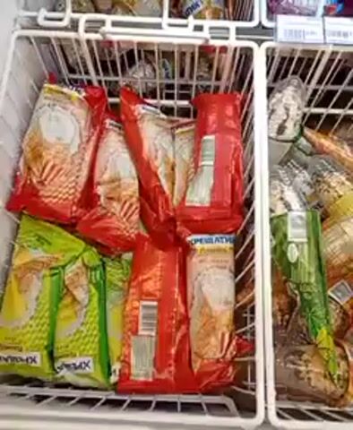 The video shows a close-up of a freezer display case filled with various frozen food items. The freezer is divided into compartments, each containing different products. In the top left compartment, there are several red-wrapped packages of what appear to be ice cream cones, some with yellow conical wrappers and others with white and red patterned wrappers. To the right of these, in the top right compartment, there are bags of popcorn and what look like frozen meat products in clear packaging. The bottom left compartment contains more red-wrapped ice cream cones, along with some green-wrapped packages and a few individually wrapped items. The bottom right compartment is filled with more ice cream cones, some with colorful wrappers depicting characters, and others with plain white and brown wrappers. The overall impression is a well-stocked freezer section of a grocery store or convenience store.