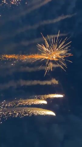 The video captures a dynamic fireworks display against a dark, cloudy night sky. Multiple fireworks launch and explode, creating bright golden and yellow sparks. Some fireworks shoot across the frame, leaving luminous trails, while others burst into starburst patterns. Several streams of golden sparks are visible, some arcing and others appearing more linear. The explosions illuminate the surrounding clouds, creating an ethereal glow. The sound accompanying the video is characterized by loud bangs and crackling, typical of fireworks. No people or text are visible in the video. The setting is at night, with discernible clouds.