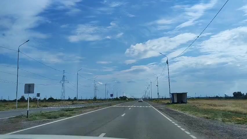 This is a forward-facing shot from a vehicle driving on a two-lane road. The road is asphalt with white lane markings and a pedestrian crossing is visible. The sky is blue with white clouds. On the left side of the road, there are fields and power lines supported by tall towers. There are also streetlights lining the road. On the right side, there are fields and a small bus stop shelter. A few cars are visible on the road ahead, and a white van passes by on the left lane.