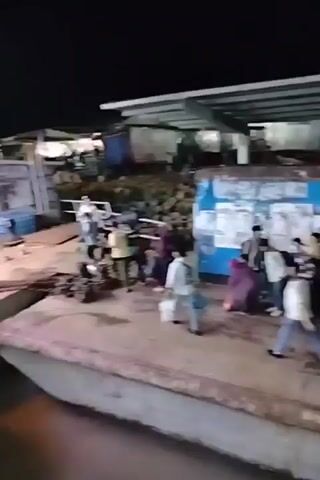 At night, many people walk away from the side of a body of water, possibly a dock. The people appear to disembark from a vessel. Some people are wearing traditional Muslim clothing while others are wearing more contemporary clothes. Some are carrying bags. Behind them is a pile of rocks, and above them is a metal canopy.