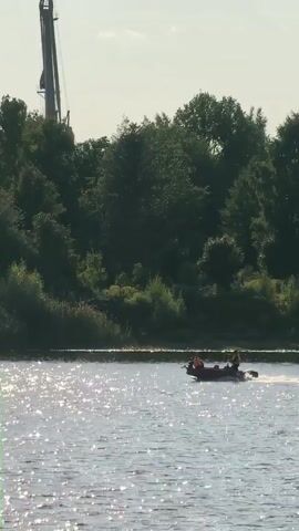 A small, dark inflatable boat with an outboard motor is navigating a body of water. Two individuals wearing orange life vests are visible inside the boat. The boat is moving from left to right across the frame. The water is choppy and reflects sunlight, creating a shimmering effect. In the background, a dense line of trees forms the shoreline. A tall, metallic structure, possibly a crane or tower, is visible rising above the trees on the right side of the frame. The sky is mostly clear with a few wispy clouds.
