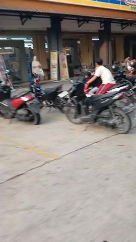 The video shows a street scene from a first-person perspective, likely from a vehicle or bicycle. The camera pans across a street lined with buildings and parked motorcycles. In the foreground, the handlebars of the vehicle/bicycle are visible. Several motorcycles are parked along the side of the road, and a few people are seen riding or interacting with them. A black car is also visible driving down the street. The environment appears to be an urban or suburban area with trees lining the street. The lighting suggests it is daytime.