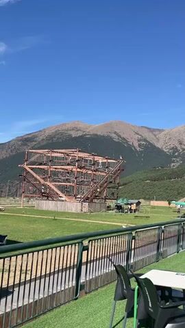 A wide shot shows a large, intricate rope course structure with multiple levels and platforms, situated in a grassy field. Behind the rope course, a mountain range with bare, rocky peaks rises against a clear blue sky dotted with white clouds and contrails. To the left of the rope course, a small wooden building is visible. In the foreground, a green railing borders a patio area with tables and chairs, some of which are occupied by people. The overall scene depicts an outdoor adventure park or recreational area with a mountainous backdrop.