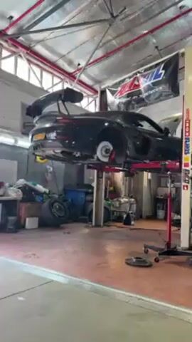 The video shows a black car on a lift in an auto repair shop. The car's wheels are off, and a man in a black shirt and cap is working on the brakes. The brake rotor is silver with numerous holes, and the brake caliper is red. The man seems to be inspecting the brake system. The shop has red brick walls, a red-painted floor, and an AMSoil banner hangs from the ceiling. There are other tools, equipment, and tires scattered around the shop.