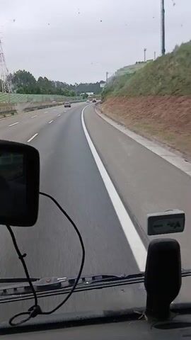 A point-of-view shot from inside a vehicle, looking forward on a highway. The road is paved with a white line marking the lane. To the right, there is a grassy slope with a concrete retaining wall. A silver device is mounted on the dashboard. Other vehicles can be seen in the distance on the highway. The sky is overcast.