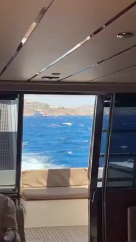 Here is a detailed description of the video:

The video shows a view from inside a boat looking out onto the ocean. The boat appears to be moving, as evidenced by the waves and white water seen through the open doorway at the rear. Visible beyond the boat are distant hills or landmasses under a bright blue sky.

Inside the boat, the interior is modern with wood flooring and padded seating. Part of a person is visible in the lower-left corner. They are wearing a light-colored shirt. The interior panels of the boat are finished in light colors with metallic trim.