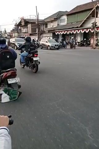The video shows a street scene with several vehicles and buildings. The perspective is from someone riding a motorcycle. In the foreground, the handlebars and a rearview mirror of a motorcycle are visible. The road appears to be paved, with white lane markings. In the background, there are various vehicles including cars, trucks, and other motorcycles. Buildings line the side of the road, some with shops or businesses. There are also power lines and utility poles. The sky is overcast, suggesting it may be daytime with cloudy weather. The traffic appears to be moderate, with vehicles moving along the road. People are visible on some of the motorcycles, wearing helmets and casual clothing. One motorcycle in the foreground has a license plate. The overall impression is a typical street scene in a populated area.