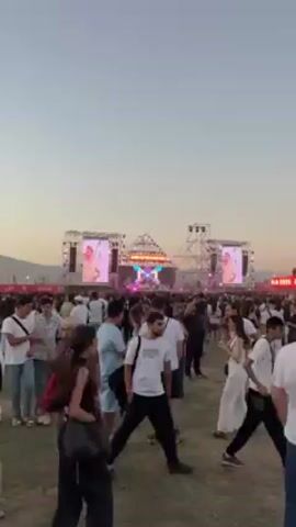 The video depicts a large outdoor music festival at dusk. The scene is dominated by a massive stage structure with large screens on either side, displaying the performing artist. There are numerous people scattered across the grass, most of them walking around or standing in groups. Many people are wearing white clothing. The sky is a pale blue, and the event is taking place on a field.