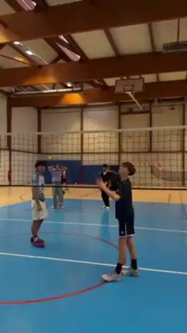 The video shows a volleyball training session inside a gymnasium with a wooden ceiling structure and skylights. The court is blue with yellow lines and a volleyball net separating the two sides. A group of young people is practicing volleyball.  

One boy, wearing a navy blue and white uniform, is about to serve the volleyball over the net. Another boy, wearing a black shirt with “SUC” printed on it and the number “7” on the back, stands ready to receive the serve. Two other boys stand further back near the net. In the background are a fire extinguisher, a blue barrier, and a soccer goal.
