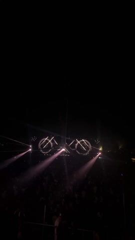 The video depicts a concert or music festival scene at night. A large crowd is visible, illuminated by spotlights. A stage is set up with a complex lighting rig consisting of geometric shapes, including circles and triangles, which are illuminated. The stage setup is relatively bright, creating a contrast with the darker crowd area.