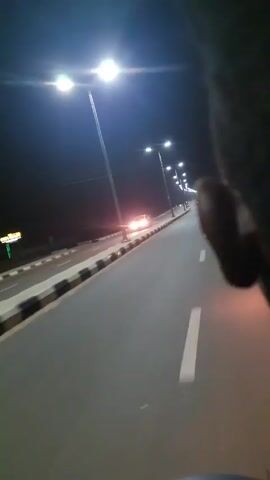 At night, a road stretches into the distance, illuminated by evenly spaced streetlights. A motorcycle and a car are visible on the road. White lane markers divide the lanes. A barrier with alternating black and white sections lines the edge of the road. The sky is dark. The camera appears to be moving forward on the road.