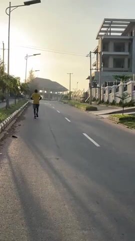 A person is jogging uphill on an asphalt road at dawn or dusk. The person is wearing a yellow shirt and black shorts. The road is bordered by sidewalks with black and white painted curbs. On the right side of the road is a large unfinished building with multiple floors and an open framework. Further up the road is a dark structure, possibly another building, partially obscured by the bright sunlight. On the left side, there are palm trees and streetlights along the sidewalk. The overall environment appears to be a residential area or a suburban street.