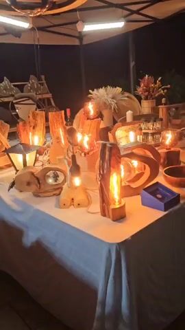The video shows a table at night, covered with a white tablecloth. On the table are various wooden lamps and decorative items. The lamps are made from pieces of wood and have Edison-style light bulbs, which are lit, giving off a warm glow. Some of the lamps are carved into unique shapes. There are also wooden bowls, platters, and other wooden crafts. In the background, there are shelves with more wooden items, some dried flowers in a vase, and a clock. The lighting suggests an outdoor market or exhibition stall. No people are visible.