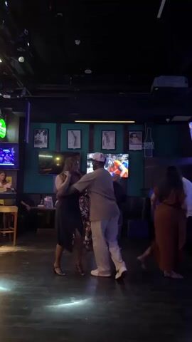 A group of people are dancing in a dimly lit room, possibly a bar or club. There are screens displaying content on the walls and a projector hanging from the ceiling. Some people are dancing in pairs, while others are dancing individually or observing. The floor is dark and reflective. The overall atmosphere appears to be lively and social.