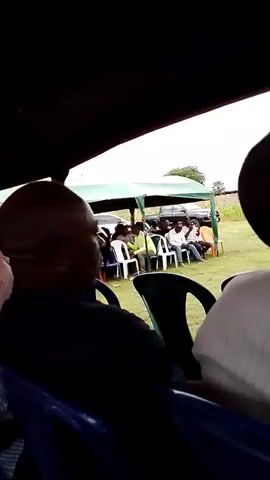 The video shows a group of people sitting under a tent. The tent has a yellow and black roof. In the foreground, a man with a shaved head and a black and white striped shirt is visible from the side. To his left, another man wearing a blue and white checkered shirt is seated. Further in the background, several other men are seated and standing. Some are wearing white shirts and others are wearing green or yellow vests. The overall impression is of an outdoor gathering or event.