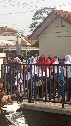 A group of young people, likely students, are gathered behind a black metal fence. Many of them are wearing headscarves, with some in red and white, suggesting a national or school-related event. They appear to be participating in some kind of competition or activity, as indicated by the cheers and counting of "juara buanya tiga" (likely meaning "the winner is three"). The environment appears to be outdoors, possibly in front of a school building, with a white building and a red-tiled roof visible in the background. A motorbike is parked in the foreground. The overall atmosphere seems festive and energetic.
