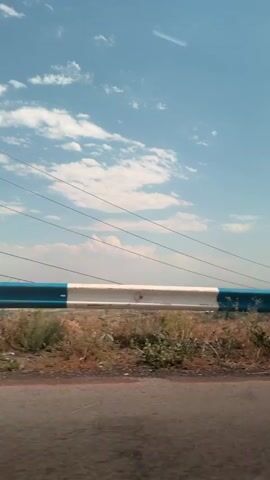 The video shows a view from a vehicle moving along a road. The camera captures a guardrail with white and blue markings, dry grass and bushes on the side of the road, and a distant landscape with a hazy horizon. The sky is blue with scattered white clouds. Power lines are visible in the background. The road surface in the foreground appears to be asphalt and shows some cracks. The scene depicts a daytime view, likely in a rural or semi-rural area, with the camera capturing a lateral motion of the landscape.