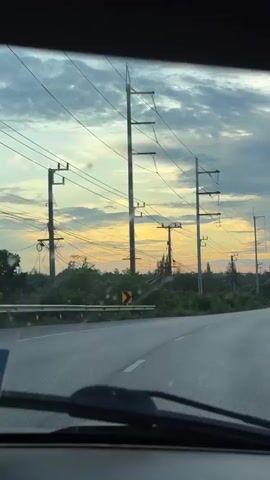 The video is shot from the perspective of someone inside a vehicle, looking forward through the windshield at a road. The road curves to the right and extends into the distance. On the left side of the road, there is lush green vegetation and several tall utility poles with numerous power lines stretching across the sky. A yellow directional sign with a black arrow indicating a curve is visible on the left side of the road. On the right side of the road, there is more vegetation, streetlights, and a guardrail. Several vehicles, including trucks and cars, are traveling on the road in the same direction as the viewpoint. The sky is filled with dramatic clouds, with a warm, orange and yellow glow from the setting sun visible on the horizon. The overall scene depicts a journey on a highway at dusk.
