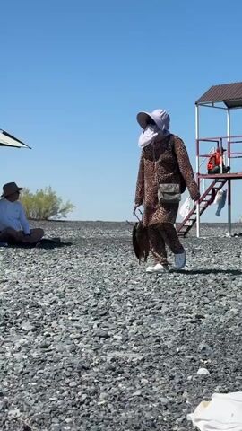 A woman wearing a leopard print outfit, a wide-brimmed hat, sunglasses, and a face covering walks across a rocky beach. She is carrying a bunch of dried fish in her right hand. A person in a straw hat sits on the beach to the left, partially obscured by an umbrella. In the background, there is a red lifeguard tower with stairs. The sky is clear and blue.