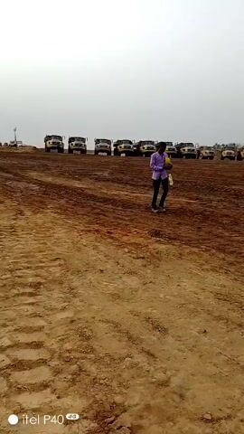 The video shows a construction or mining site on an overcast day. A line of large yellow dump trucks are parked in the background. In the foreground, the ground is dirt and appears to have been recently worked. Several people wearing casual clothes or safety vests are visible, some walking, some standing together. A tall communication tower is visible in the distance, as well as some temporary buildings. The video quality is somewhat poor.