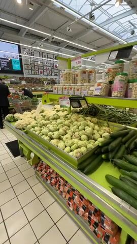 A low-angle shot pans across a brightly lit supermarket produce section. In the foreground, a shopping cart filled with groceries is visible. The camera then moves to focus on displays of fresh vegetables, including cauliflowers and turnips, with price tags indicating 1.49 and 7.99. Further back, two men wearing dark clothing are seen in the produce section, one pushing a shopping cart. Overhead, digital screens display advertisements for produce and meat products. The environment is clean with tiled flooring and fluorescent lighting.