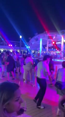A group of people are dancing on a deck at night. The deck is illuminated by colored lights, including blue, pink, and red. In the background, there is a stage with a band or DJ setup. People are dressed in casual summer attire. Some individuals are holding drinks. The overall atmosphere appears to be a party or celebration.