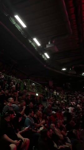 The video shows a large crowd of people seated in what appears to be an indoor arena or stadium. The lighting is dim, with overhead fluorescent lights interspersed with red lighting, creating a dark, reddish ambiance. The camera pans across the crowd, showing a diverse mix of people, some seated and others standing or walking. A large video screen is visible in the distance, displaying images of people. The structure of the building is visible above, showing metal beams and ductwork. The overall impression is of a crowded event, possibly a sporting event, concert, or convention.