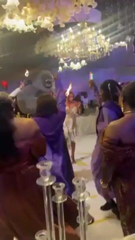 A group of people are gathered in an ornately decorated room, illuminated by chandeliers and colored lights. A woman wearing a white dress stands in the center of the room. Near her, someone in a gorilla costume is illuminated by blue lights. Other people are holding torches. The crowd appears to be enjoying a celebration.
