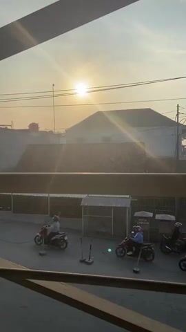 This video captures a street scene at sunrise in Wirosari, Indonesia. The low-angle sun casts a warm, hazy glow over the landscape, with visible lens flares creating a dreamy effect. The sky is bright and clear, suggesting fair weather.

The primary subjects are several motorcycles with riders, navigating a paved road. In the foreground, a horizontal wooden railing frames the view, suggesting the footage is taken from an elevated position, perhaps a balcony or window. In the mid-ground, there are a few small, simple structures, one of which appears to be a covered stall or kiosk. A fence runs along the back of this area. Beyond this, residential or commercial buildings are visible, with their roofs and upper levels silhouetted against the bright sky.

The activity appears to be the start of the day, with people commuting or going about their morning routines on their motorcycles. The overall mood is tranquil and serene, enhanced by the soft morning light and the quiet movement of the vehicles.

Notable details include the presence of what looks like a water tower in the distance on the left. On one of the structures in the mid-ground, there is visible text on a sign that reads "KOTAK LAMARAN," which translates to "Application Box" in Indonesian, possibly indicating a job application drop-off point or a similar administrative function. The riders are mostly wearing helmets, a common safety practice. The image conveys a sense of everyday life beginning in a tropical or sub-tropical setting.
