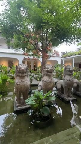 The video begins by showing a courtyard with a pond. Several statues of mythical creatures, possibly lions or gargoyles, are submerged in the pond. Lush green plants are growing in and around the pond, with some plants floating on the surface. In the background, a white building with an arcade is visible. The building is adorned with lights that create a warm glow. A large tree with hanging lights is in the center of the courtyard. The camera pans down to the pond, where a small, bright red fish is swimming. The reflection of the surrounding trees and the building can be seen on the surface of the water. The video ends with a close-up of the water and the fish.