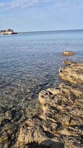 A still video shows a rocky coastline meeting the clear blue ocean. The water is calm with gentle ripples. The rocks are light brown and tan, some are partially submerged. The sky is a pale blue with a few wispy clouds. In the distance, there are faint shapes of buoys or possibly boats. A small building or structure is visible on the land to the far left.